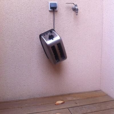 A toaster hanging from a plug on a rosa wall. A bread in front of it on the floor.